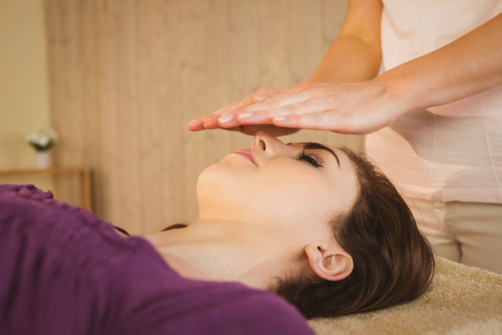 Woman laying on a massage table with hands hovering over her face-Third Eye Expansion Massage