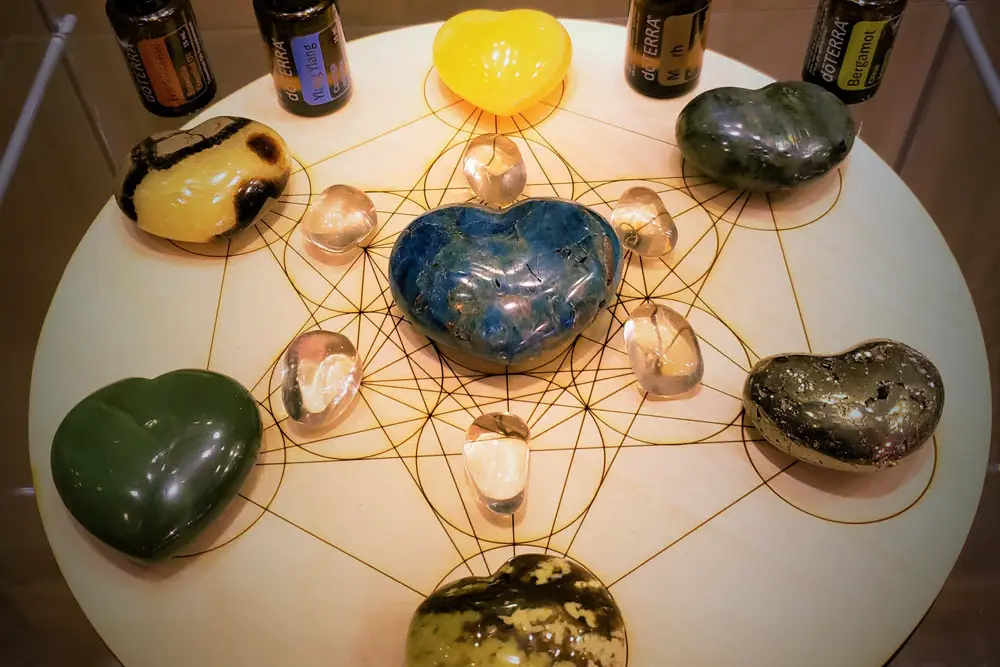 A sacred geometry crystal grid with a large blue heart-shaped stone centered on a Flower of Life pattern, surrounded by smaller stones and essential oils for a wellness session.