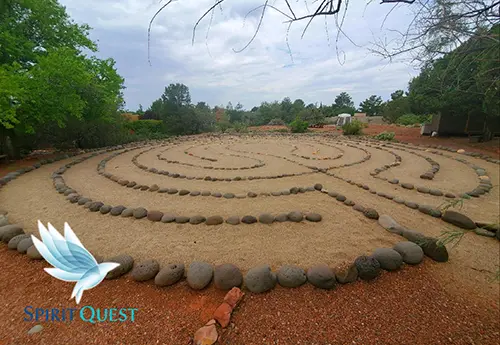 A stone walking labyrinth laid out on the red dirt ground in Sedona, Arizona, surrounded by green desert vegetation and trees under soft daylight.